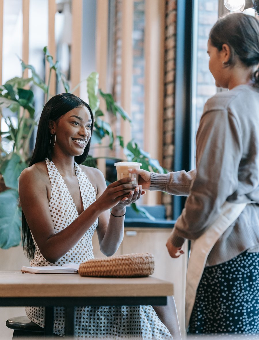 Cliente sorrindo sentada à mesa de uma cafeteria, recebendo o copo das mãos da atendente.